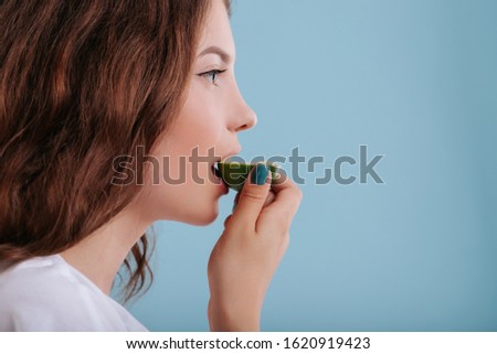 girl eats slice of lime, make grimaces, in studio, isolated on blue background, copy space, close up, profile view