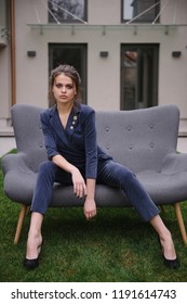 Girl In Elegant Blue Costume Posing On Grey Sofa Outside. Seductive Elegant Woman Sitting On Coach. Fashion Portrait Of Brunette Stylish Female Model In Arm Chair