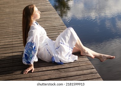 a girl in an embroidered Ukrainian shirt sits on the pier, the reflection of clouds in the water of the lake. On the shore of the sky. vyshyvanka day. freedom. patriot. - Shutterstock ID 2181599913
