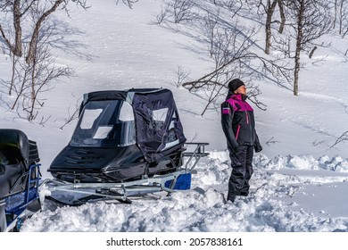 Girl Enjoing Strong Sun At Mountain Slope Beside Snowmobile With Sled After Long Drive. Much Snow In Scandinavian Mountains, Holidays In Lappland, Northern Sweden