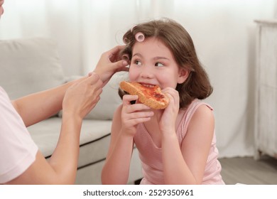 Girl enjoys eating toast while mother fixes her hair with curlers. The scene captures a lighthearted bonding moment between them, blending beauty routine and casual breakfast time. - Powered by Shutterstock