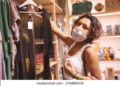 Girl With A Face Mask Choosing Clothes Inside A Shopping Mall. Selective Focus. Concept Of New Normality And Safe Shopping.