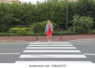A girl with fair hair in a red dress and a denim jacket with a bag on her shoulder crosses the road at a pedestrian crossing. The concept of traffic rules