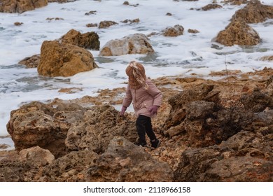 A Girl In A Fashionable Knitted Balaclava Walks On The Shore Of The Winter Sea. Child Wearing A Jacket