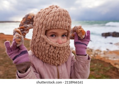 A Girl In A Fashionable Knitted Balaclava Walks On The Shore Of The Winter Sea. Child Wearing A Jacket