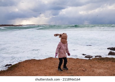 A Girl In A Fashionable Knitted Balaclava Walks On The Shore Of The Winter Sea. Child Wearing A Jacket