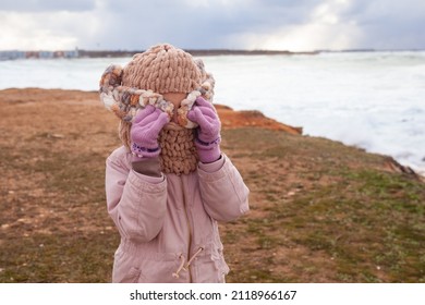 A Girl In A Fashionable Knitted Balaclava Walks On The Shore Of The Winter Sea. Child Wearing A Jacket