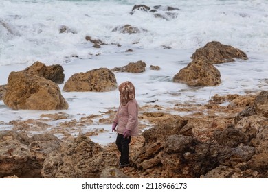 A Girl In A Fashionable Knitted Balaclava Walks On The Shore Of The Winter Sea. Child Wearing A Jacket