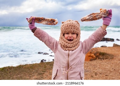 A Girl In A Fashionable Knitted Balaclava Walks On The Shore Of The Winter Sea. Child Wearing A Jacket