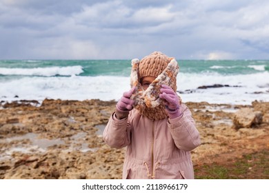 A Girl In A Fashionable Knitted Balaclava Walks On The Shore Of The Winter Sea. Child Wearing A Jacket