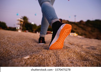 Girl Feet In Sneakers With Bright Orange Soles Go Away On The Sand. Concept For Start, Going, Step Forward, Fashion.