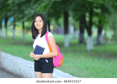 Girl Female Teenager With Black Hair In A Bob White East Asian Woman Green Park With A Backpack Background. Lady Walking, Goes, Walk Away, Look At The Camera.