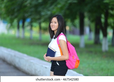 Girl Female Teenager With Black Hair In A Bob White East Asian Woman Green Park With A Backpack Background. Lady Walking, Goes, Walk Away, Look At The Camera.