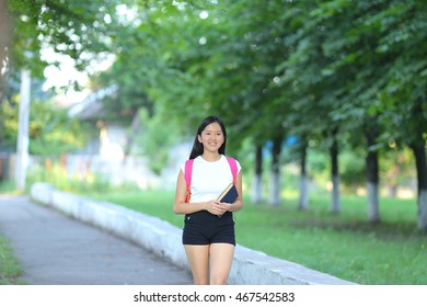 Girl Female Teenager With Black Hair In A Bob White East Asian Woman Green Park With A Backpack Background. Lady Walking, Goes, Walk Away, Look At The Camera.