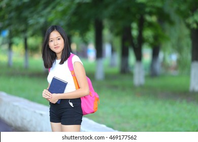 Girl Female Teenager With Black Hair In A Bob White East Asian Woman Green Park With A Backpack Background. Lady Walking, Goes, Walk Away, Look At The Camera.