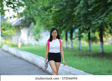 Girl Female Teenager With Black Hair In A Bob White East Asian Woman Green Park With A Backpack Background. Lady Walking, Goes, Walk Away, Look At The Camera.