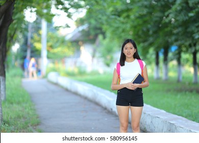 Girl Female Teenager With Black Hair In A Bob White East Asian Woman Green Park With A Backpack Background. Lady Walking, Goes, Walk Away, Look At The Camera.