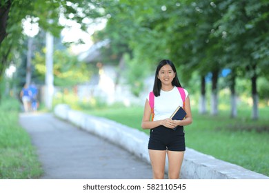 Girl Female Teenager With Black Hair In A Bob White East Asian Woman Green Park With A Backpack Background. Lady Walking, Goes, Walk Away, Look At The Camera.