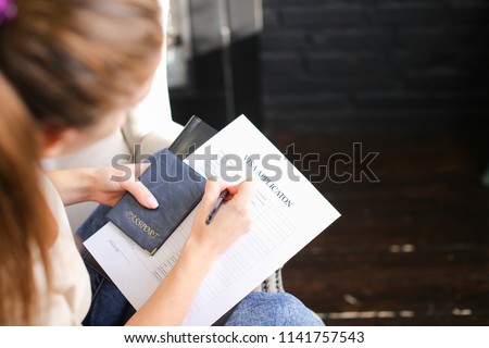 girl filling visa application form, student sitting on grey chair with passport. Young fair-haired woman wearing beige T-shirt jeans holding planchette with papers writing. Concept of execution of