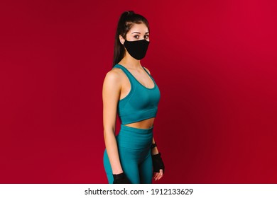 Girl fitness trainer in a protective mask dressed in sportswear looks at the camera. Studio photo on a red background with space on the side - Shutterstock ID 1912133629