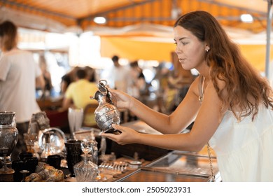 Girl in flea market store look at silver decanter, antique gifts. Female buyer holds item in hands and examines product, makes purchase decision - Powered by Shutterstock