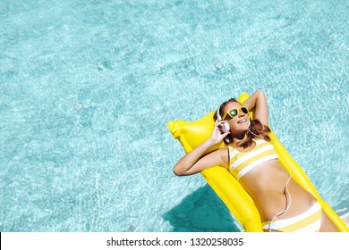 Girl Floating On Yellow Beach Mattress And Listening To Music In Earphones In The Blue Pool. Summer Holiday Idyllic Concept Top View.