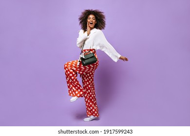 Girl with fluffy hair in white blouse posing on lilac backdrop. Stylish lady in red polka dot trousers with handbag smiling on isolated background.. - Powered by Shutterstock