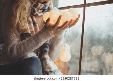 A girl in a fluffy sweater is resting by the window with a festive garland in her hands. Christmas mood, winter, cozy holidays, fairy tale atmosphere. Glowing garland balls.