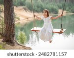 girl in the forest swinging on a swing. Rope swing on a forest lake. Barefoot girl in a white dress with long hair.