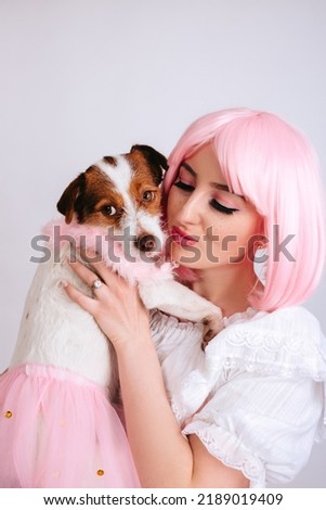 A girl with freckles and pink hair, with a bright pink make-up, hugs a dog in pink clothes on a white background. Close-up portrait of a girl with a pet dressed in pink Halloween costumes.