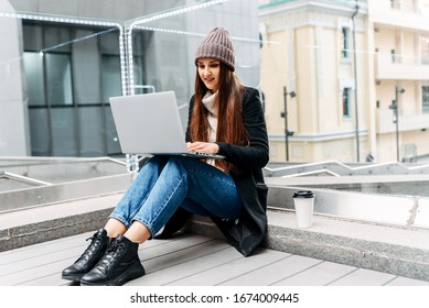 Girl freelancer with a laptop works sitting on a step on the street. She is in casual clothes - Shutterstock ID 1674009445