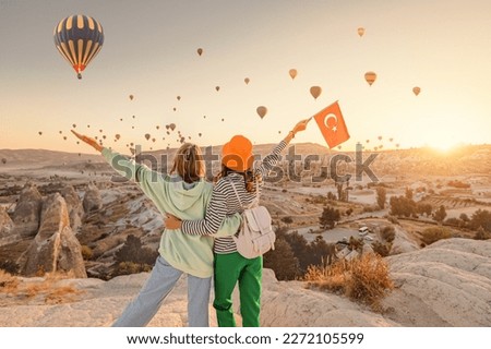 Girl friends travellers with turkish flag, hugging on a viewpoint and admiring view of flying hot air balloons in Cappadocia