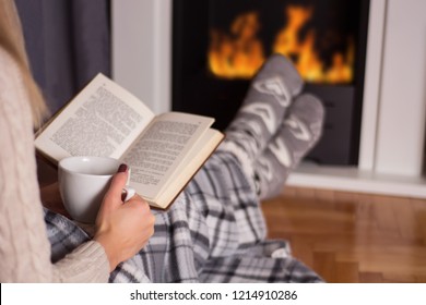 Girl in front of the fireplace reading book and warming feet on fire and legs are covered with blanket, in hand holds cup of hot tea. Winter and cold weather concept at home. Close up, selective focus - Powered by Shutterstock