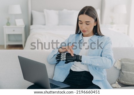 Girl in front of pc is setting bionic prosthesis. Young european woman is adjusting artificial hand. Attractive women is working at home. Modern bionic prosthesis.
