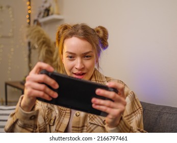 Girl gamer enthusiastically plays a video game on a portable game console at home, in the living room. Room interior. Beige tones. Cyberspace, esports, online games with friends.