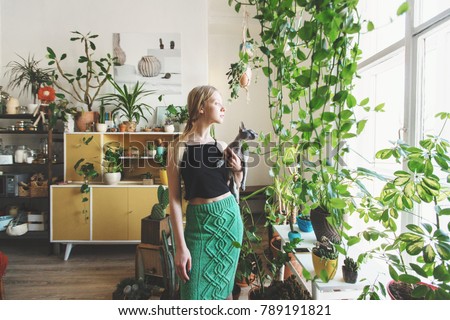 girl gardener with a cat in her arms looking out the window against a background of indoor plants