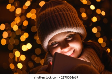 Girl And Gift Box With Christmas Lights. Smiling Girl In A Hat, Scarf Looking At The Camera At Night With Hard Wrong Light, Close Up. Holidays Theme