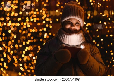 Girl And Gift Box With Christmas Lights. Smiling Girl In A Hat, Scarf Looking At The Camera At Night With Hard Wrong Light, Close Up. Holidays Theme