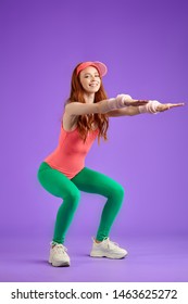 Girl With Ginger Hair Doing Warm Up Exercises Active Workout Fitness Trainer In Gym, Dressed In Aerobics Clothes, Makes Squats And Keeps Hands Forward, Looks In Camera With Smile. 80s Style Concept