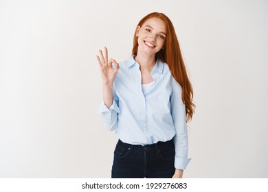 Girl With Ginger Hair Showing OK Sign And Smiling, Recommending Your Promotion Product, Standing Satisfied Against White Background.