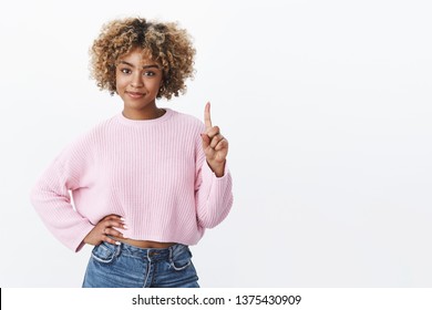 Girl giving directions smiling and looking confident at camera holding hand on waist showing number one with finger and smirking assertive ordering coffee posing over white background upbeat