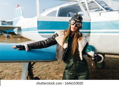 a girl with glasses and a headdress holds a helmet in her hands, in the background of the plane she is dressed in a warm leather jacket, gloves, green overalls