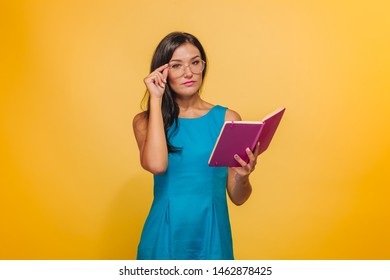 Girl with glasses on a yellow background in a blue dress reading a pink notebook - Shutterstock ID 1462878425