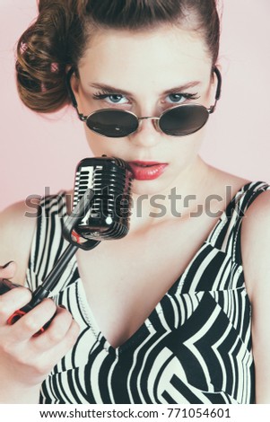 Girl in glasses sing in microphone. Pin up young girl on pink background, radio. Music, look and retro style, pinup. Beauty and vintage fashion. Woman singer with stylish retro hair and makeup.
