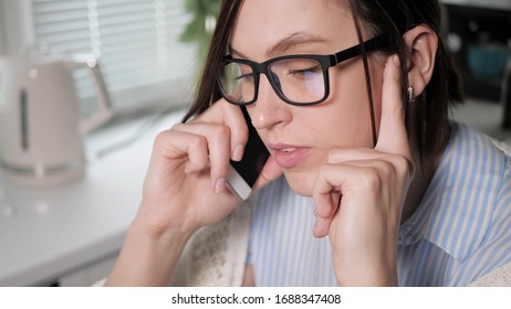 Girl With Glasses Talking Intently On Phone In Kitchen, Holding Her Head Hands Screen Is Reflected In Lenses. Girl Freelancer At Work, Difficult Conversation Finding Out Personal Relationships Concept