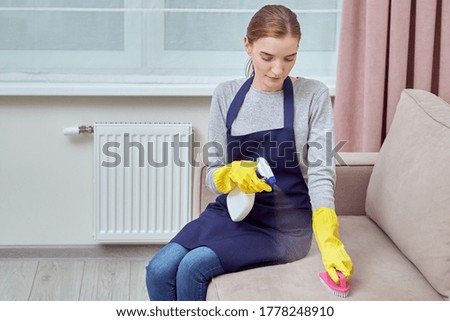 A girl in gloves cleans upholstered furniture with brush and household chemicals