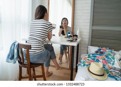 Girl Going On Vacation Get Ready In Bedroom Sitting In Front Mirror. Young Asian Woman In Casual Wear Wake Up Early Doing Make Up In Dressing Table Looking At Herself With Cosmetics In Cozy Apartment