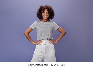 Girl in gray t-shirt is smiling on isolated background. Dark-skinned curly woman in white shorts laughing and posing on purple backdrop - Powered by Shutterstock