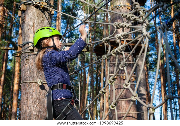a girl in a green helmet with a\
safety net, smiling, runs along a rope wall made of ropes in the\
form of a cobweb a distance in a forest extreme rope\
park