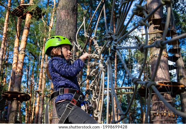 a girl in a green helmet with a\
safety net, smiling, runs along a rope wall made of ropes in the\
form of a cobweb a distance in a forest extreme rope\
park